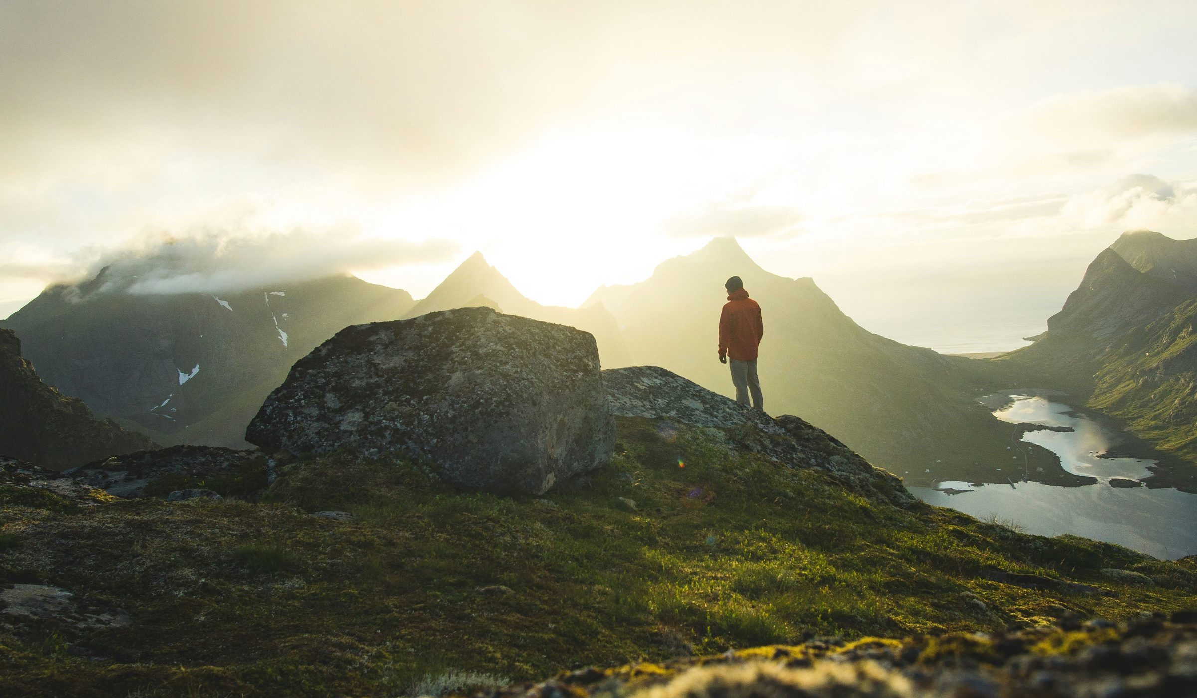 A walker standing on a grass ridge at sunrise, looking out over mountains and a fjord.